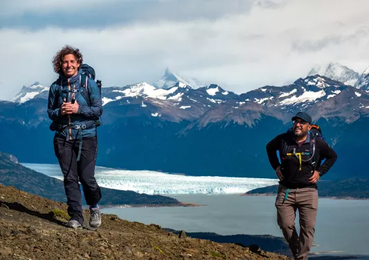 Two guests on craggy hillside, lake and snowy mountains in background.