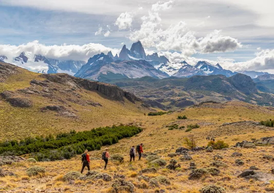 Four guests trekking over grassy, rocky meadow, large snowy mountains in background.