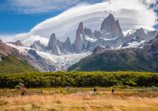 Three guests walking in grassland, forest, craggy mountains, clouds behind them.