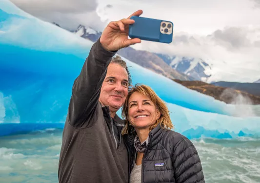 Two guests taking selfie in front of large glacier.