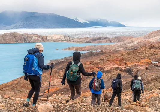 Six guests hiking down stony cliffside, blue water, mountains visible in distance.