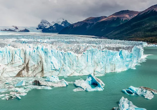 Wide shot of large glacial shelf, dark clouds and mountains in background.