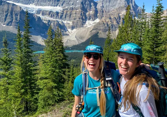 Two guests smiling for camera, snowy mountain, trees, blue sky in background.