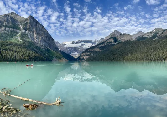 Wide shot of large blue lake, people canoeing in distance, mountains, clouds, sky in background.