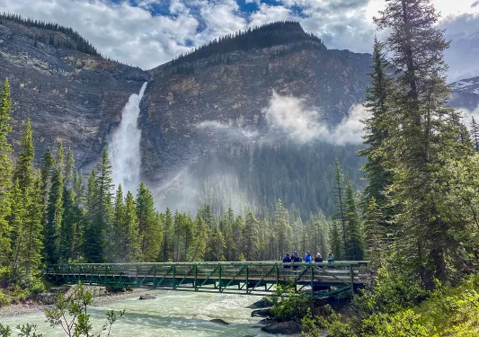 Wide shot of guests crossing bridge over blue river, mountains in background.