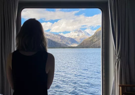 Guest on cruise ship in room, looking out towards mountains.