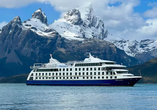 Wide shot of cruise ship, craggy mountains, clouds.