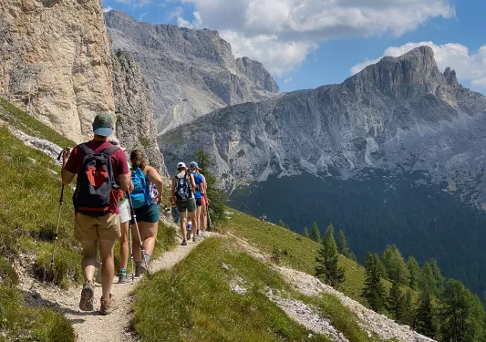 Group of guests walking along hillside trail, cliffs to left, sprawling mountains to right.