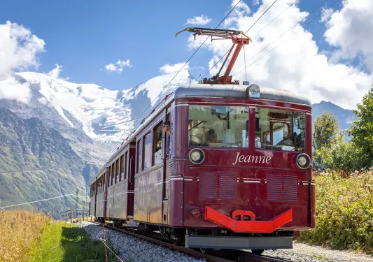 Shot of a red train, "Jeanne", mountains & clouds behind it.