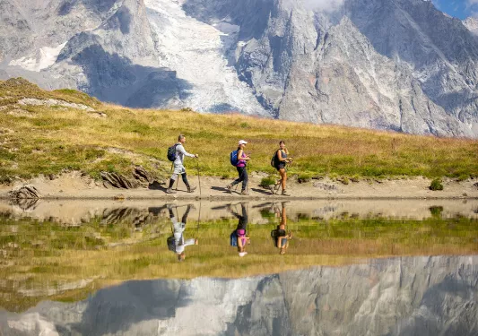 Three guests walking past reflective lake, meadow, snowy mountain in distance. 