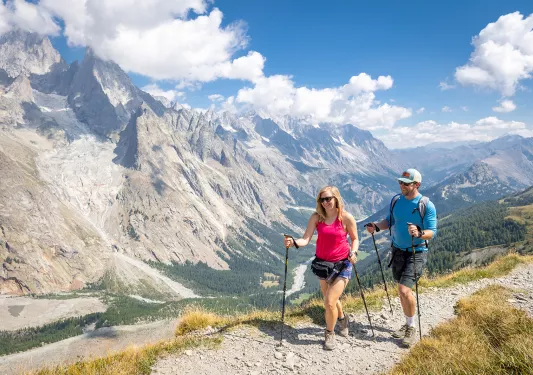 Two guests hiking on mountaintop trail, large mountain vista in background.