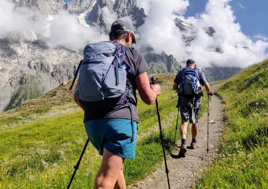 Two guests walking up mountain trail, clouds, mountain in distance. 