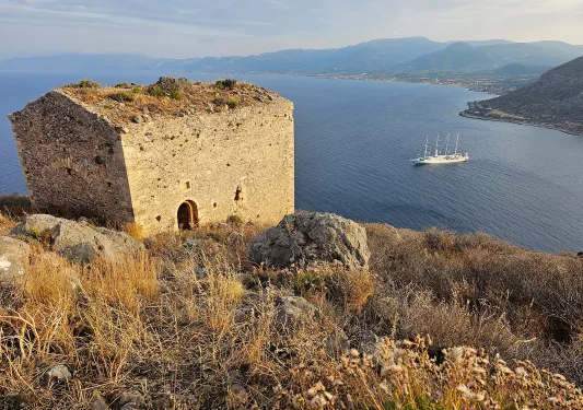 Shot of small stone shack on cliffside, ocean, mountainous coast, boat behind it.