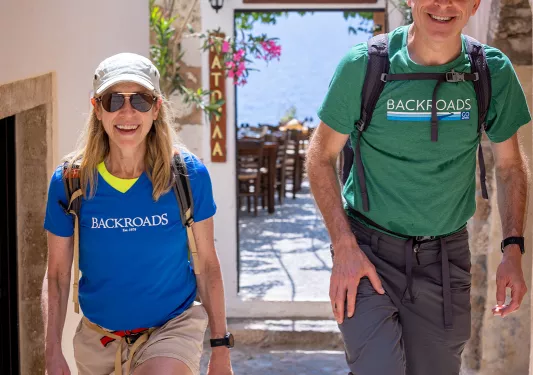 Two guests walking down narrow Grecian alleyway towards camera.