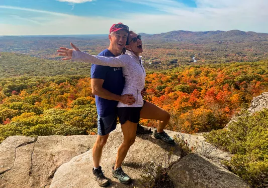 Two guests on clifftop, red/yellow/orange trees below.