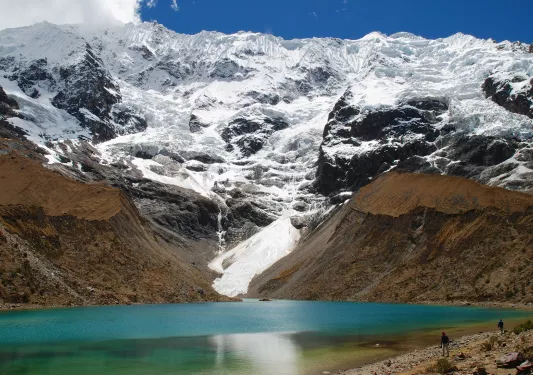 Wide shot of blue lake, brown rock, snowy mountains in distance. 