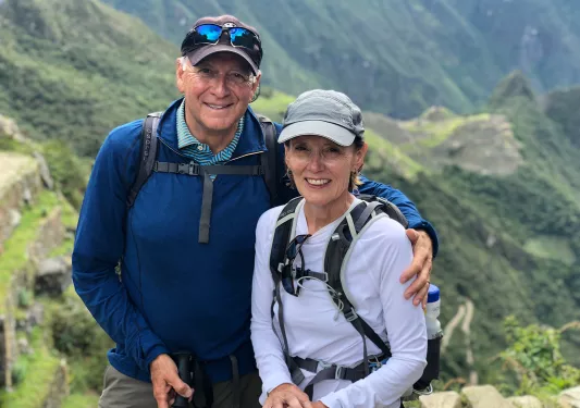 two guests embracing and smiling for camera, large grassy hills behind them.