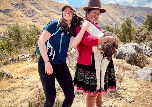 Guest, local woman and baby llama standing together. Llama sniffing guest.