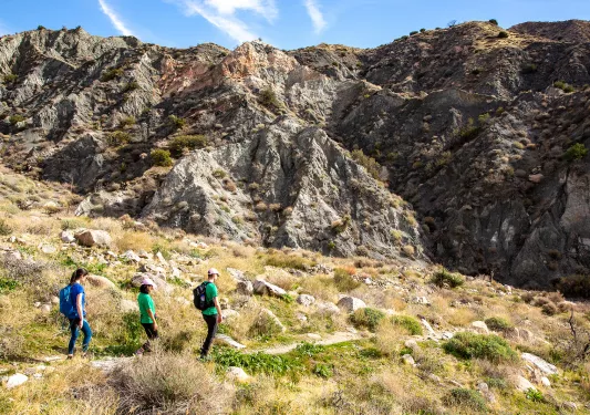 Group of three hiking in desert, craggy hill in background.