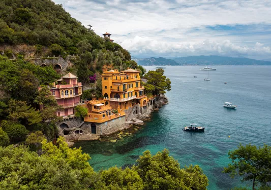 Wide shot of coastal town, two buildings on hill, one red, one gold. Ships in water below.