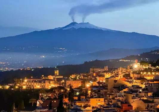 Nighttime shot of Italian hillside town, smoking volcanoes in distance.