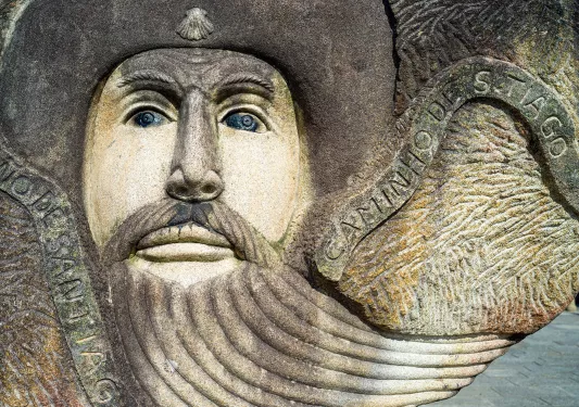 Shot of stone sculpted face, "CAMINO DE SANTIAGO" beside it.