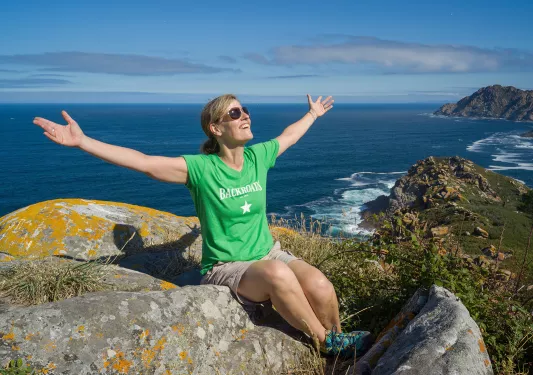 Guest on sunny hilltop, arms outstretched, ocean behind them.