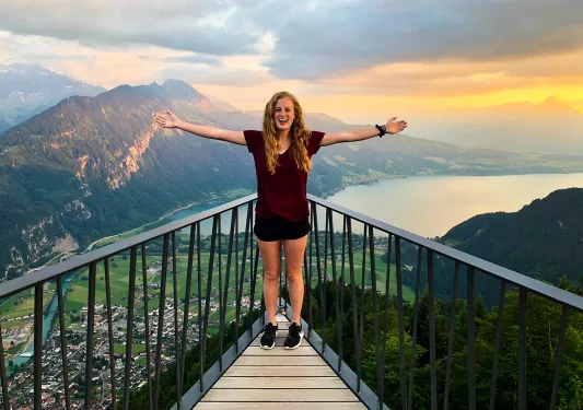 Young guest above mountain town, cliffs, lake, clouds in distance. 