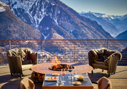 Wooden outdoor patio with wooden chairs around a wood firepit, and large mountains in the background