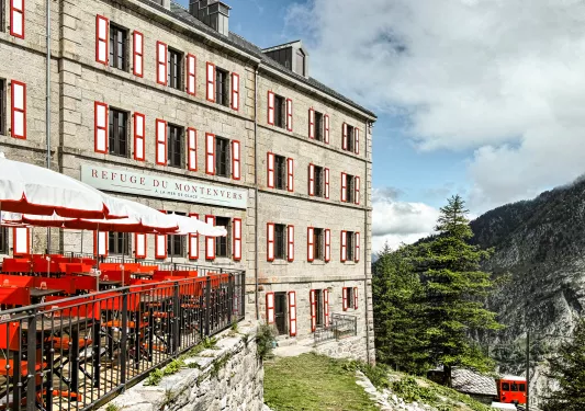 Stone hotel building exterior, with a metal railed balcony with red and it chairs and umbrellas