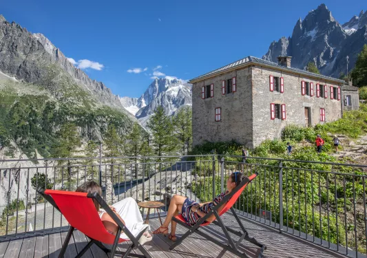 Two women sitting on reclining chairs on a wooden balcony, looking out to large mountains