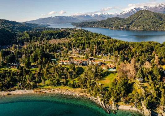 Sky view of white and brown buildings surrounded by a forest and a lake in the distance