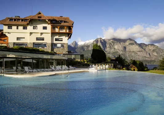 Outdoor pool with white chairs and a white building and large mountains in the background