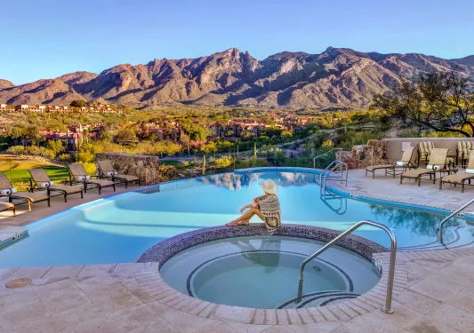 Woman sitting on stone in between a pool and hut tub, looking out towards tall mountains