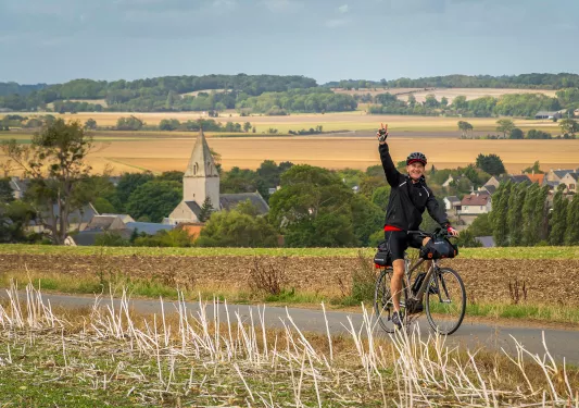 Man biking on an asphalt road in a grass valley, while he's waving his hand in the air