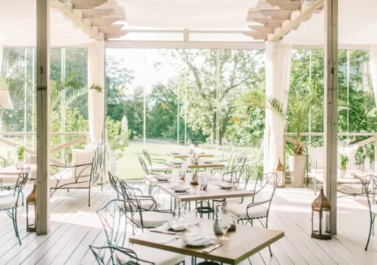 Indoor dining area with white chairs and wooden tables, with large glass walls surrounding