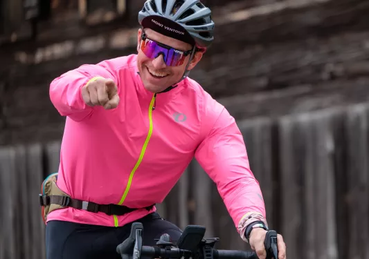 Man with a pink jacket riding a bicycle and pointing at the camera