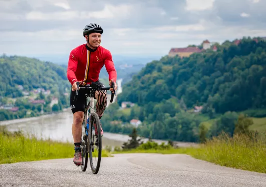 Man riding a bike on an asphalt road, with mountains of trees behind him