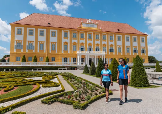 Two women laughing and walking in an outdoor garden in front of a yellow and white building
