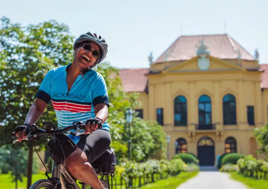 Woman wearing sunglasses and riding a bike in front of a long pathway leading to a yellow building