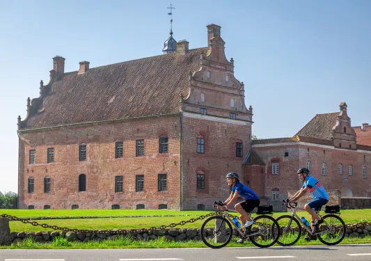 Two bikers in front of a large red building in the middle of a grass field