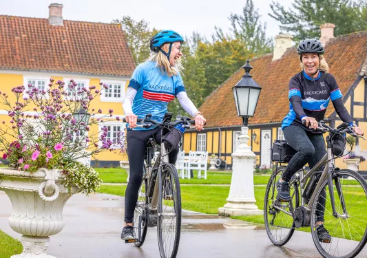 Two bikers smiling with on a road with yellow buildings in the background