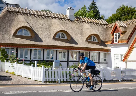 Man biking with a house and a white picket fence behind him