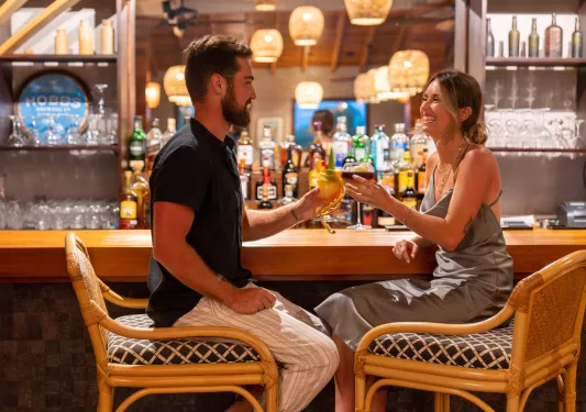Man and woman sitting in front of a bar, raising glasses of beer and wine