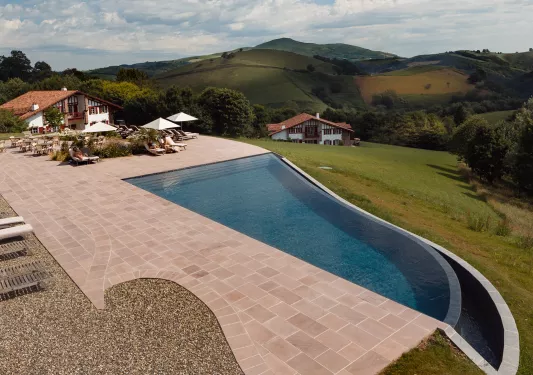 Outdoor pool with brick walkway to the left and pool chairs by a building
