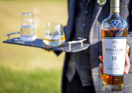 Person in a suit holding a tray of glasses and a bottle of Scotch whisky