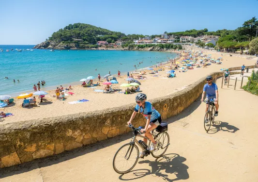 Man and woman biking on a stone pathway, with a small beach filled with crowds of people behind them
