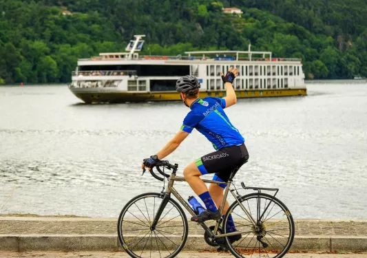 Man in a blue jersey, biking on a road next to a lake while waking at a boat