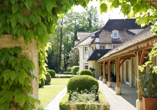 Outdoor view of white and brown building with bushes and tall trees in the background