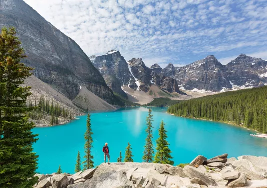 Man standing on a boulder, looking out to a blue lake and tall trees